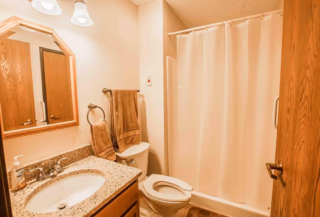 A bathroom with a granite countertop sink, wooden framed mirror, toilet, beige shower curtain, and brown towels hanging on a towel rack and ring.