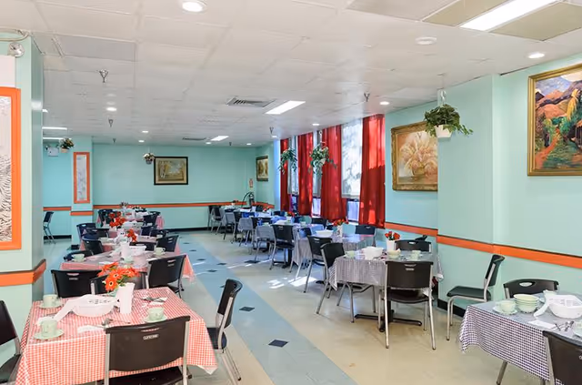 Communal dining room with checkered-tablecloth tables set with dishes, chairs, red curtains and framed paintings.