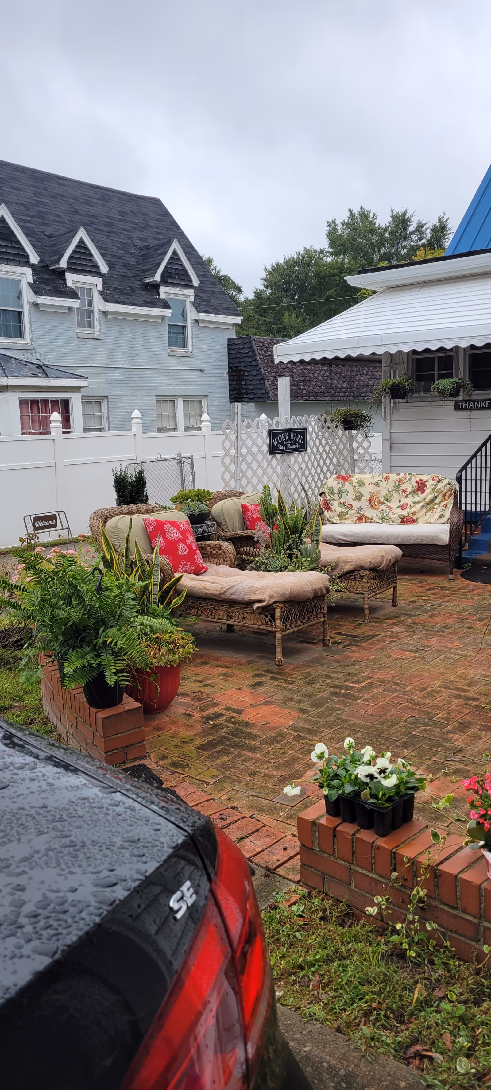 Outdoor brick patio with wicker lounge chairs, potted plants, and a bench beside a white fence.