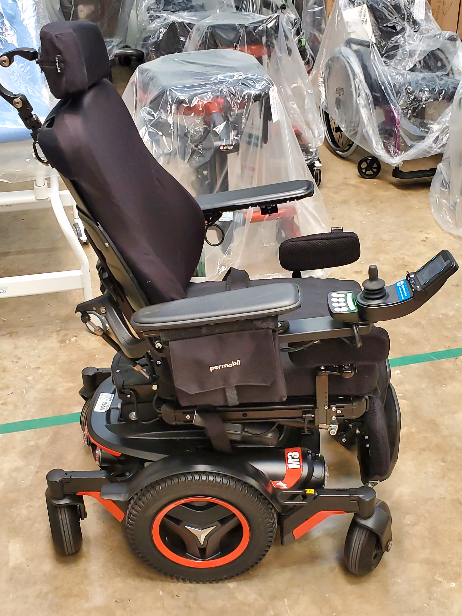 An electric wheelchair sits in an indoor storage area with plastic-covered equipment.