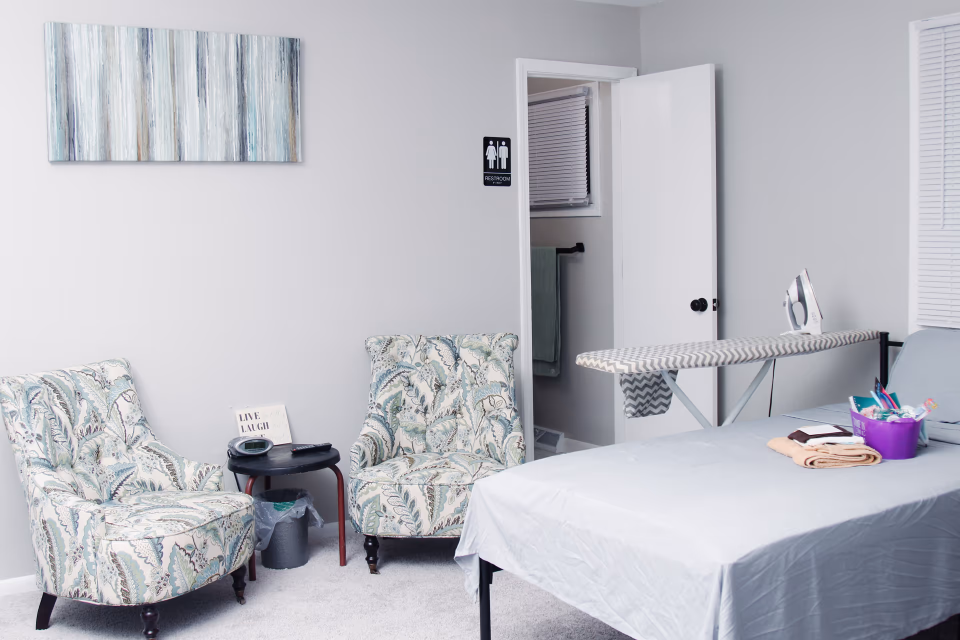 Small, light-gray room with two patterned armchairs, a small table, a covered bed, and an ironing board with an iron.