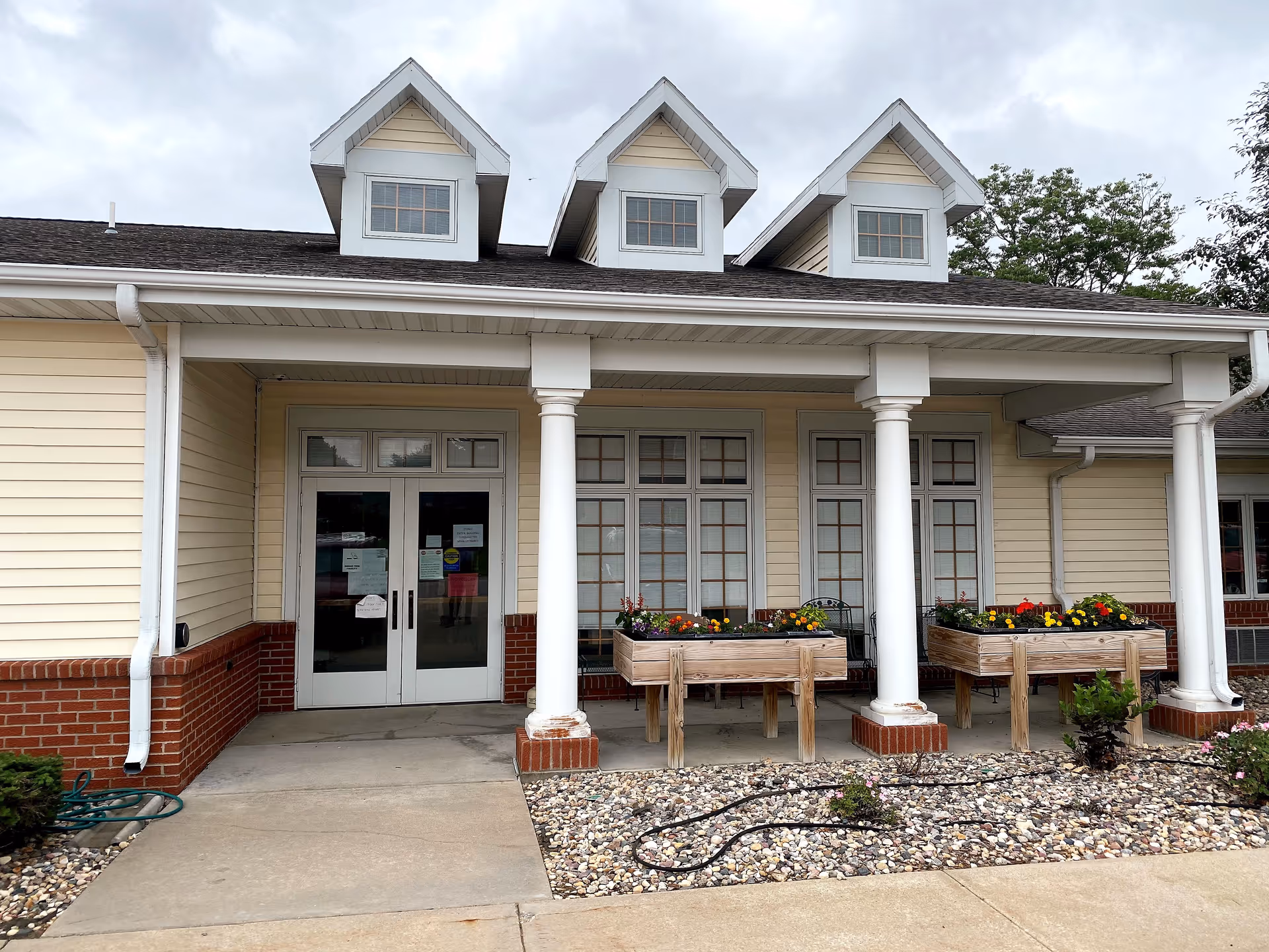 Front entrance of a beige senior living facility with white columns and flower planters.