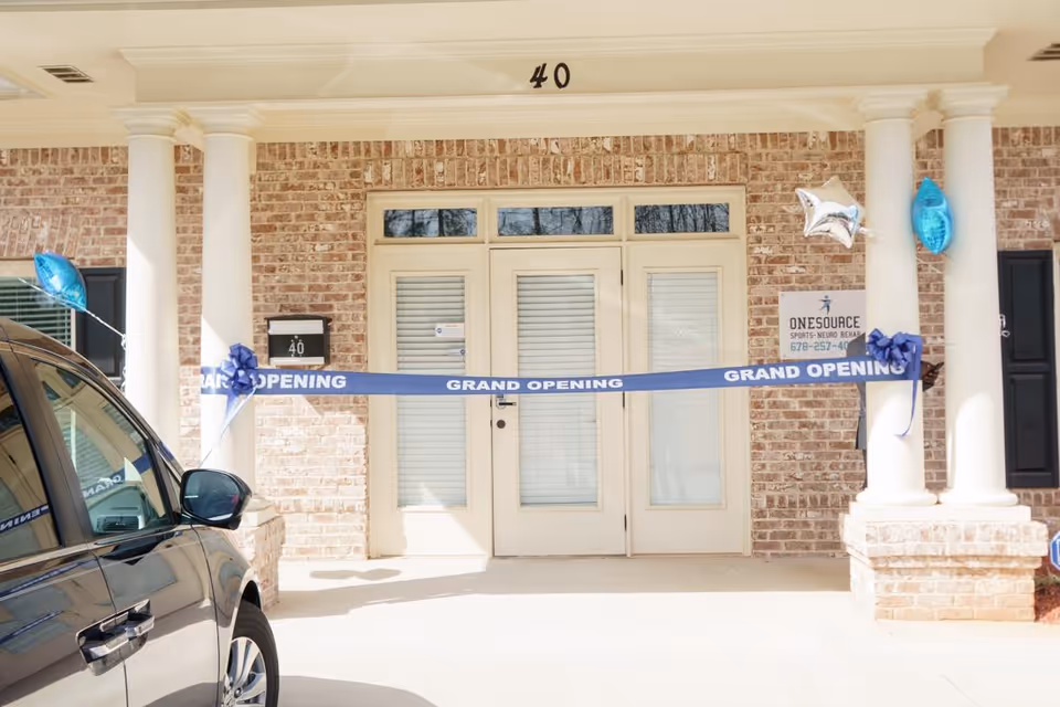 Brick front entrance with grand opening ribbon, blue balloons, and a parked car nearby.