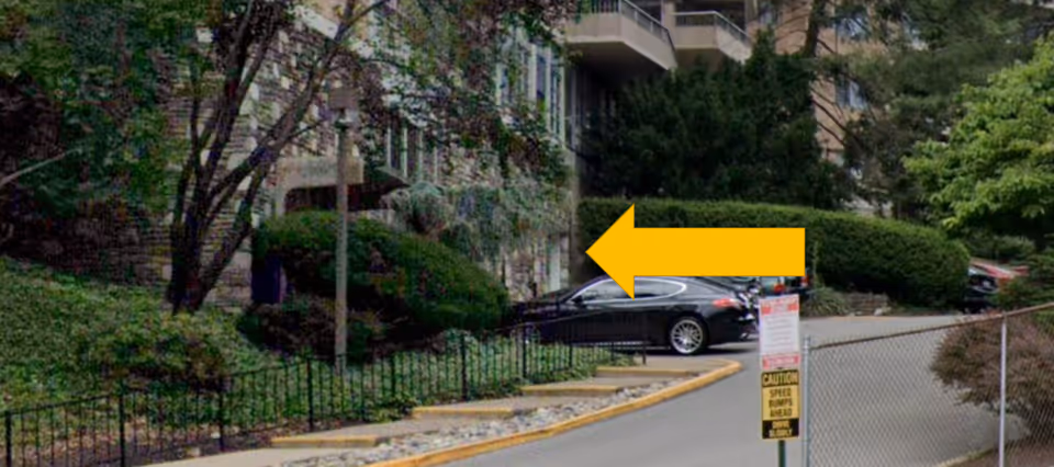 Exterior view of a senior living building entrance with driveway, yellow arrow overlay, cars, and greenery.
