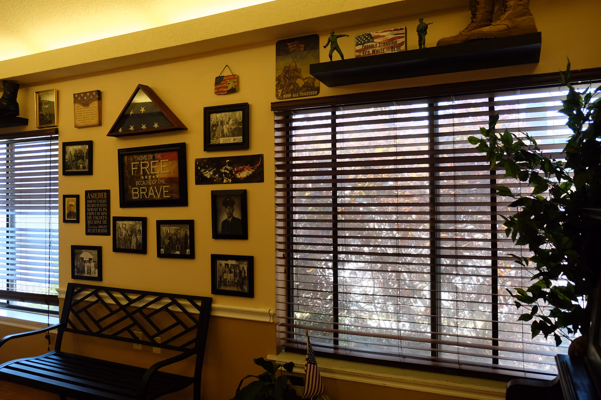 Interior lounge with a bench, a wall of framed patriotic photos and plaques, and large blinds-covered windows next to a potted plant.
