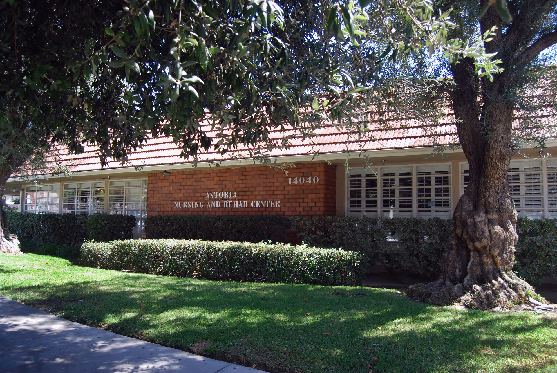 Exterior view of Astoria Nursing and Rehab Center building with a brick facade, large windows with white shutters, surrounded by green bushes and trees, and a well-maintained lawn in front.