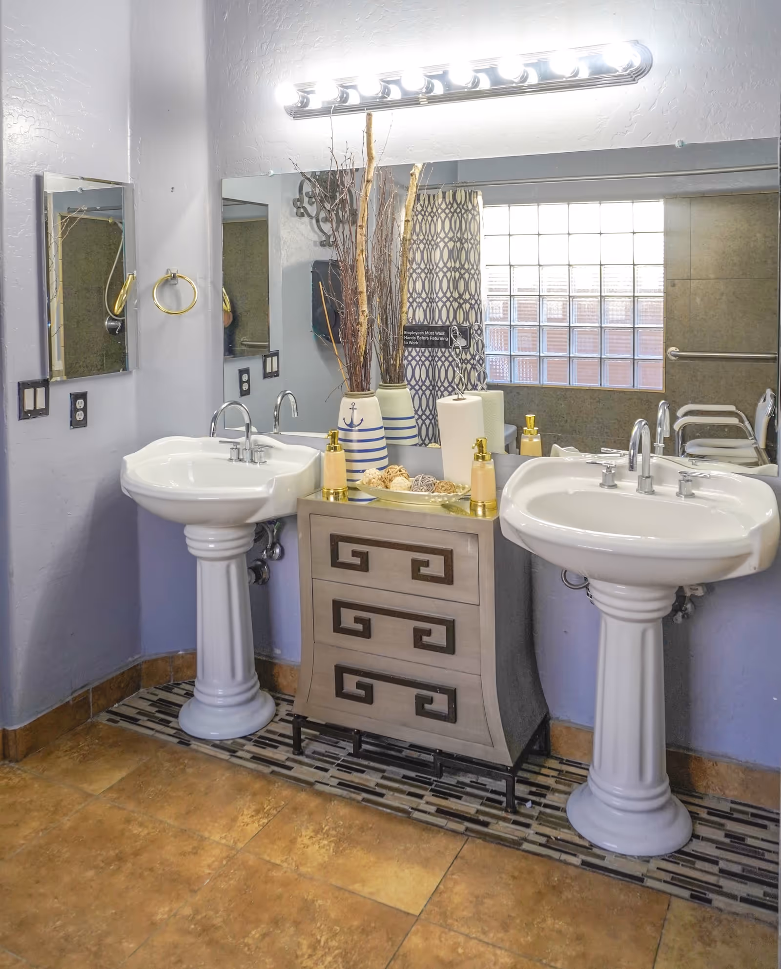 Two pedestal sinks flank a decorative cabinet in a bright shared bathroom.
