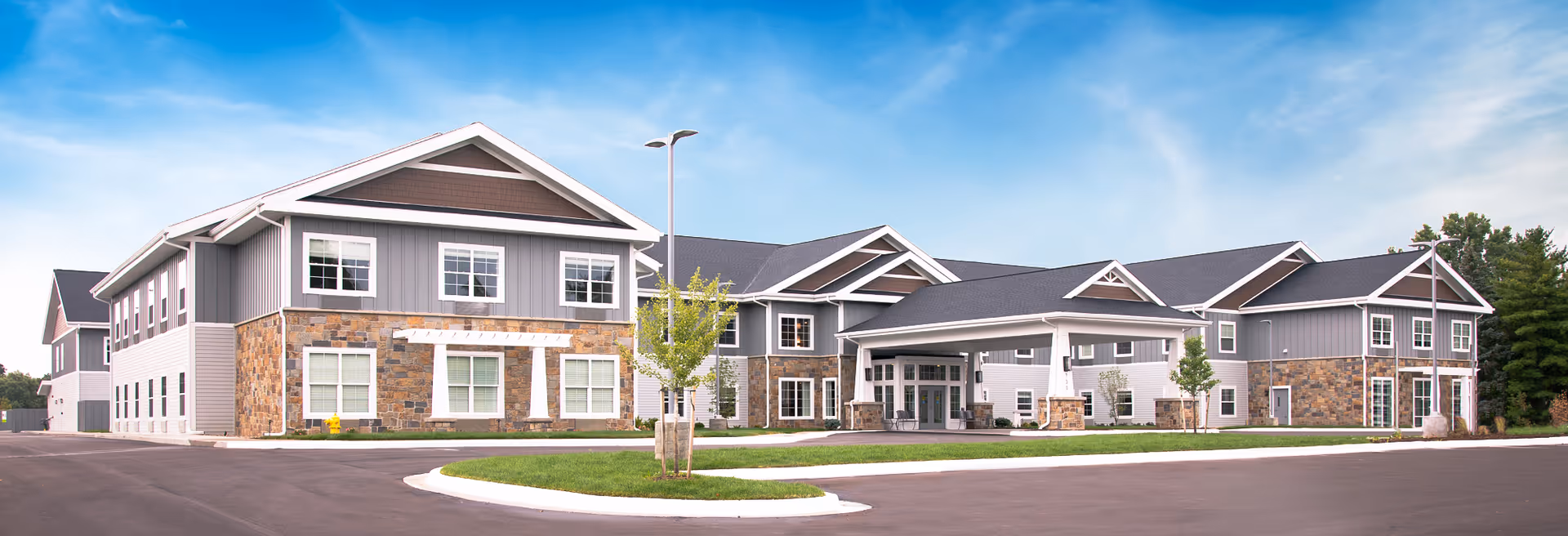 Exterior front view of a modern two-story senior living facility with a covered main entrance, stone and siding facade, and landscaped driveway.