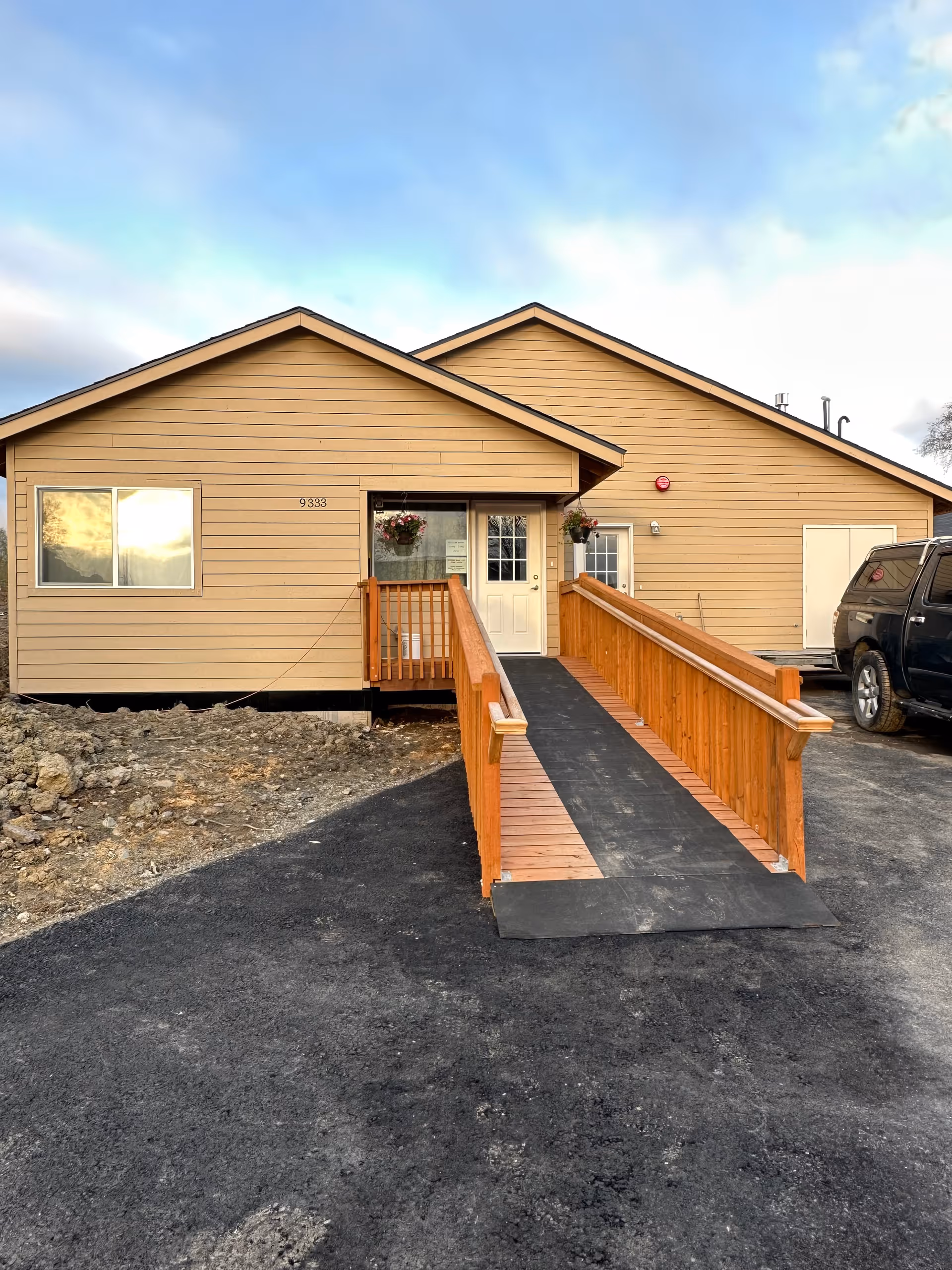 Single-story beige building with wooden accessibility ramp and hanging flower baskets at entrance.