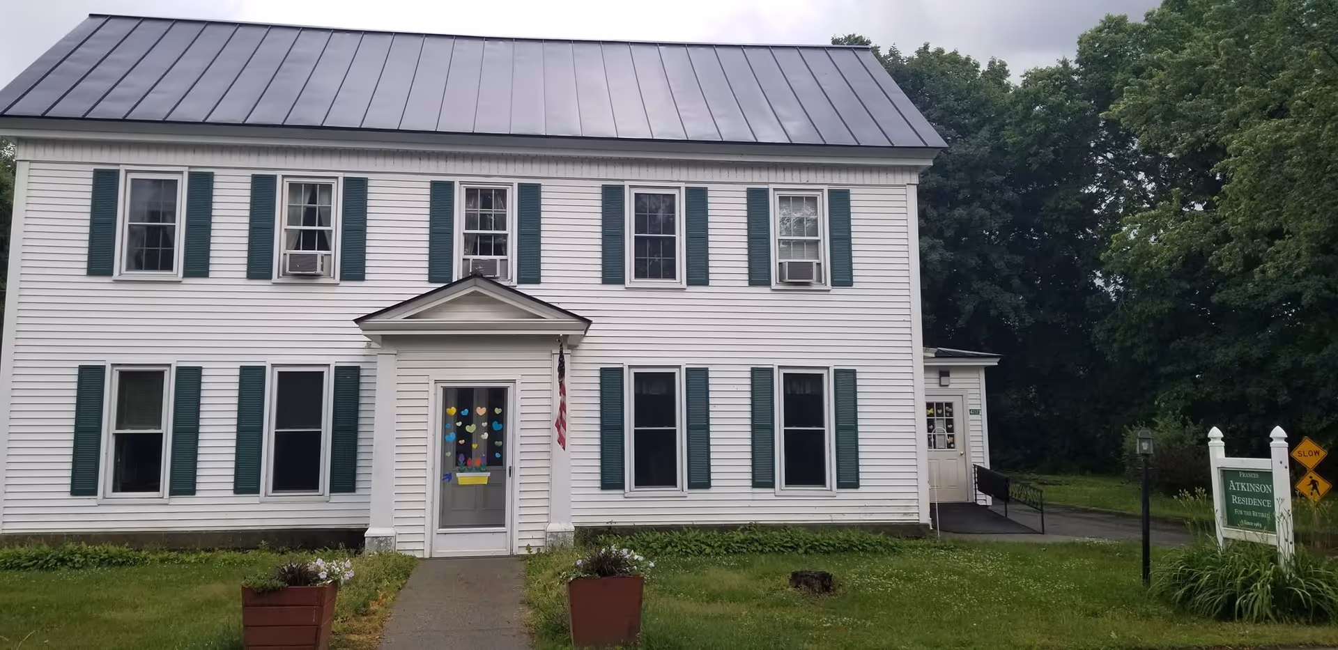 Front exterior view of a two-story white building with green shutters and a metal roof. The entrance door has colorful heart-shaped decorations. There is an American flag hanging beside the door. A sign on the lawn reads 'Frances Atkinson Residence For The Retired'. The building is surrounded by grass and trees.