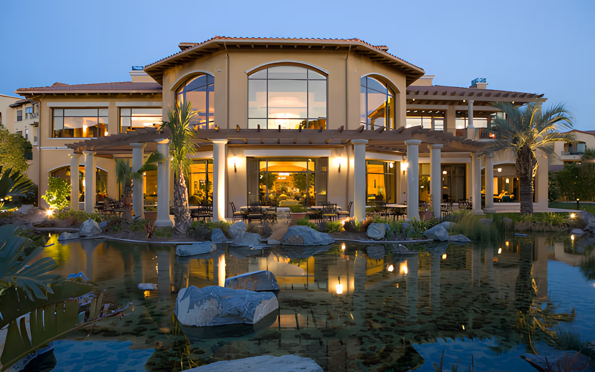 Well-lit two-story senior living building with large arched windows, a columned outdoor patio, and a reflecting pond in front at dusk.