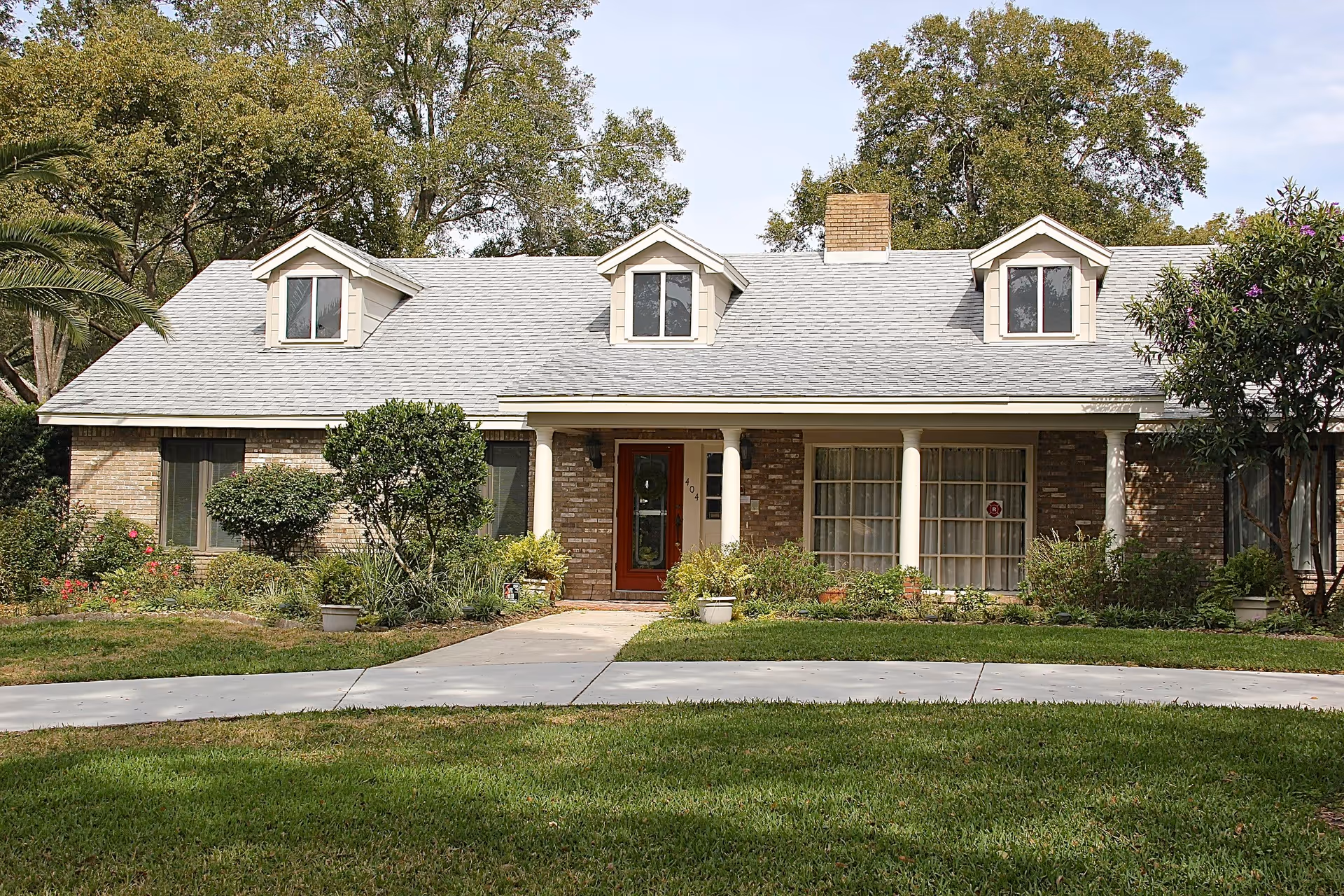 Front exterior view of a single-story brick house with a gray shingled roof, three dormer windows, a chimney, and a red front door. The house is surrounded by green grass, shrubs, and trees under a clear sky.