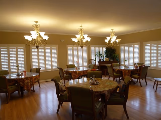 Bright senior living dining room with round tables, floral cloths, chandeliers, shutters.