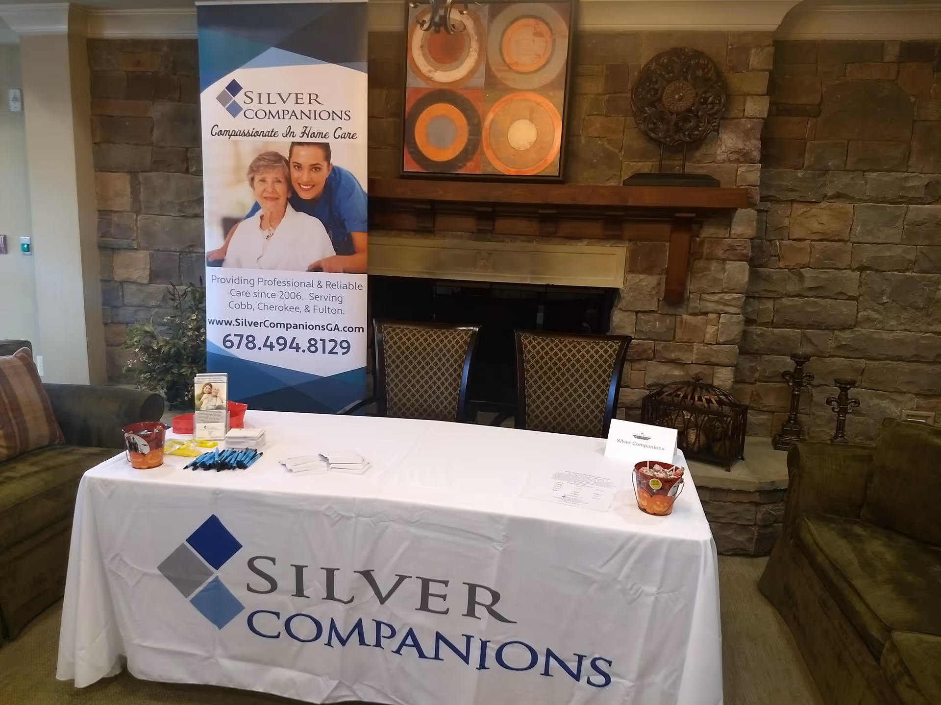 Indoor lounge area with a promotional table and banner for Silver Companions near a stone fireplace.