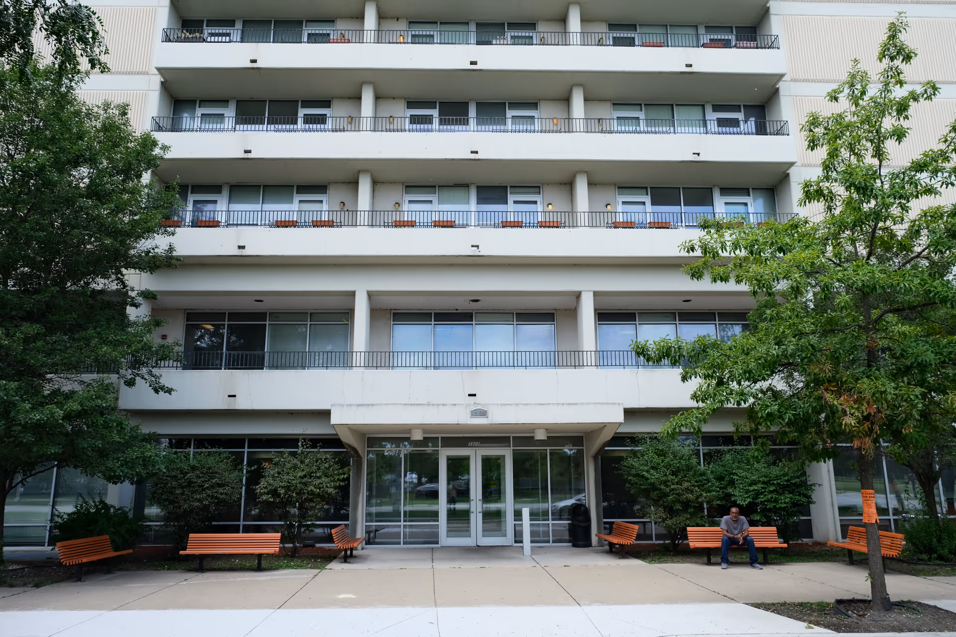 Front entrance of a multi-story building with balconies, trees, orange benches, and a person sitting on a bench.