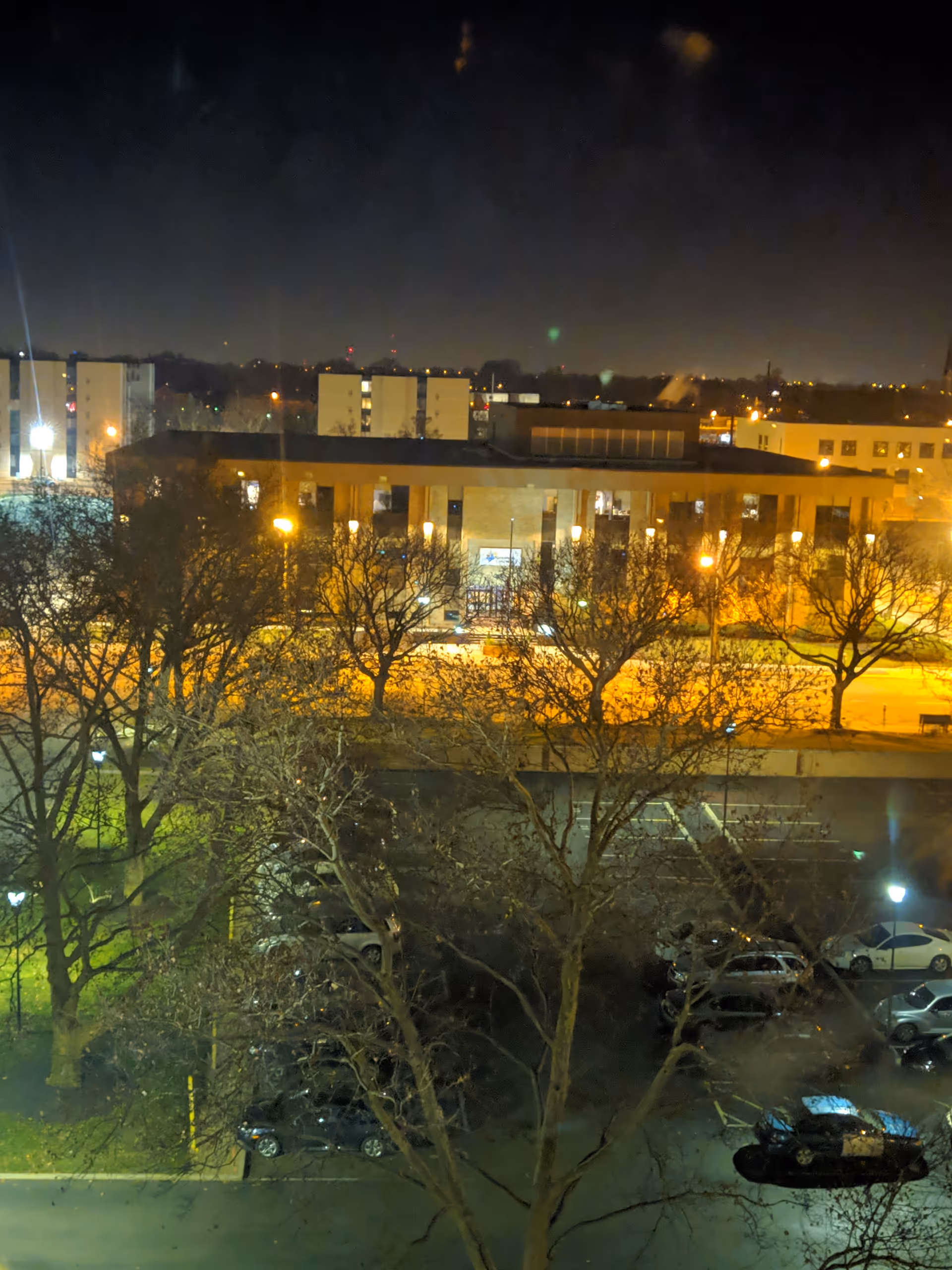 Nighttime exterior view of a building and parking lot with leafless trees.