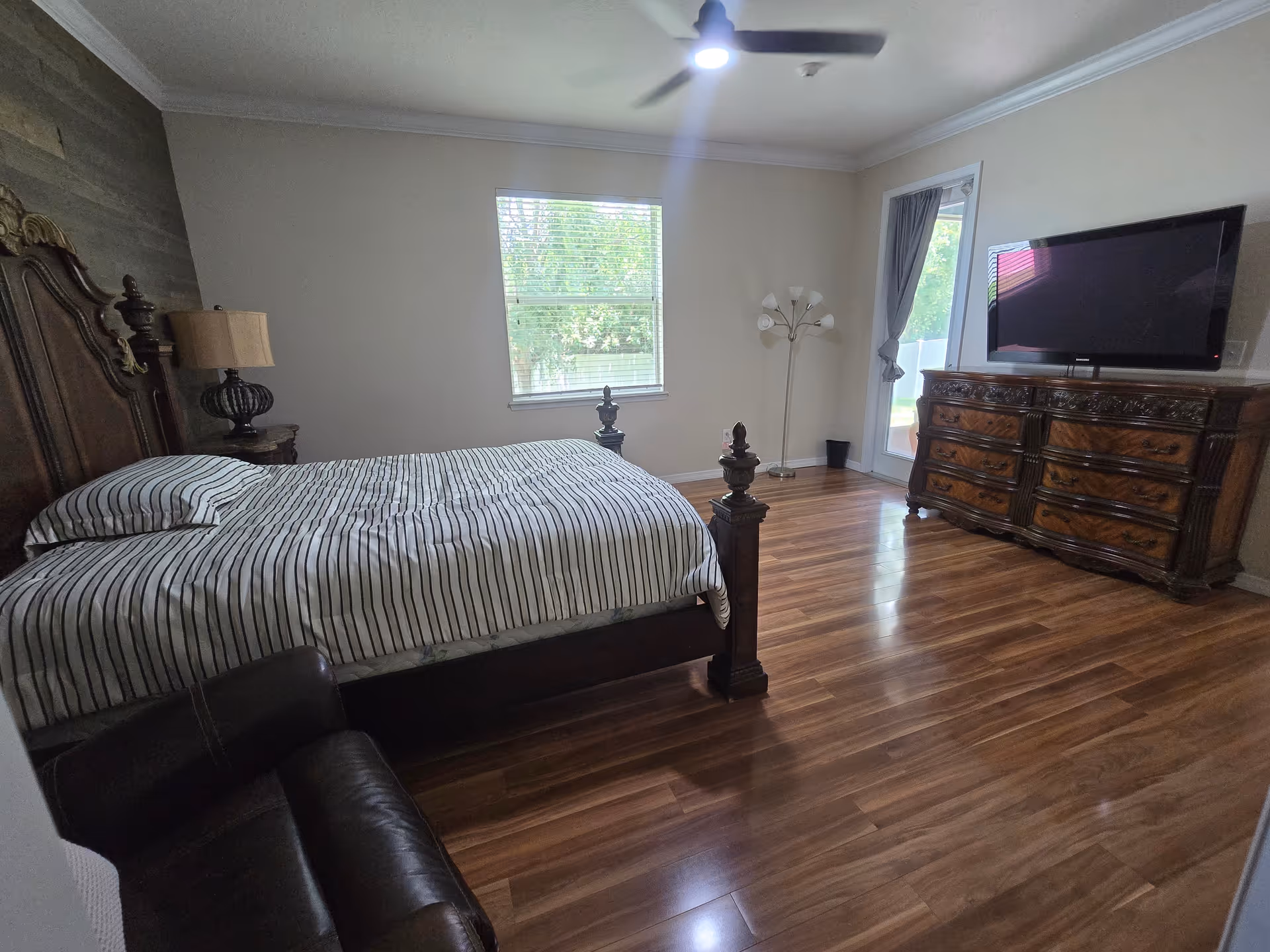 A spacious bedroom with a wooden bed, dresser, TV, and large windows.