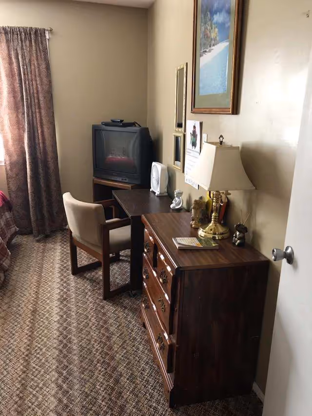 Senior living room featuring a desk, dresser, lamp, TV, chair, and framed art.