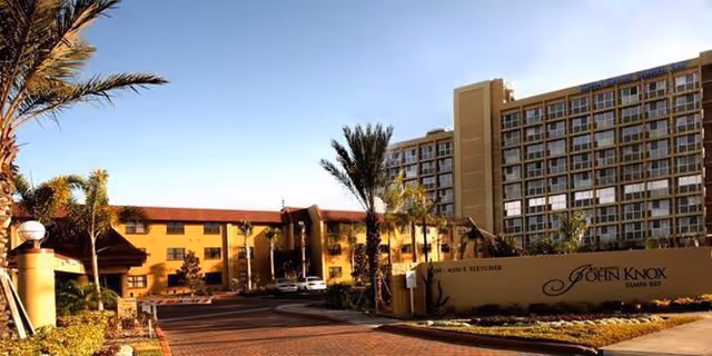 Front entrance of a multi-story senior living complex with palm trees, a driveway, and a sign reading 'John Knox Tampa Bay'.