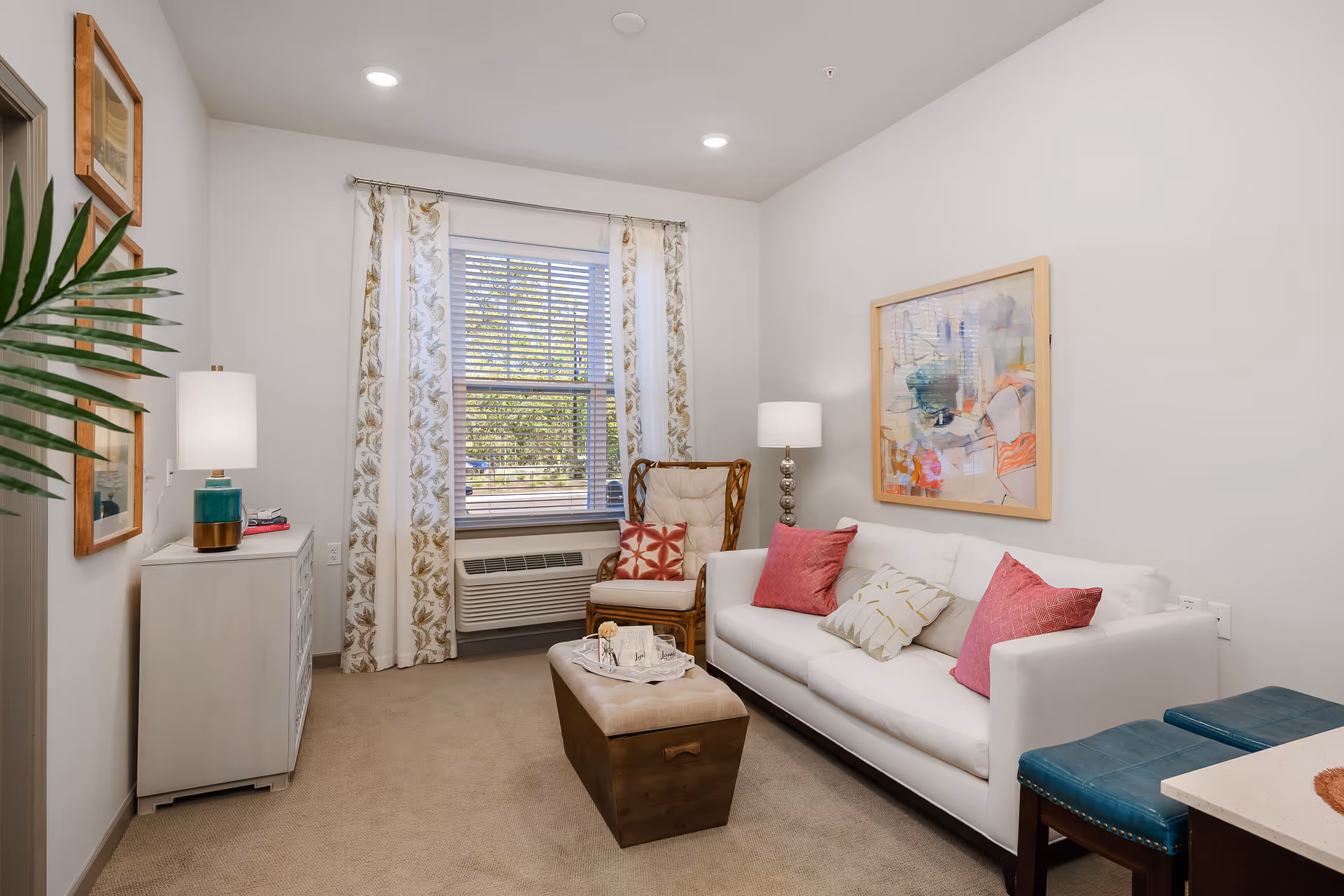 A cozy living room with a white sofa adorned with pink and patterned pillows, a wooden armchair with a cushion, a small ottoman coffee table, a white dresser with a lamp, and a window with floral curtains letting in natural light. The walls are decorated with framed artwork and the room has beige carpeting.
