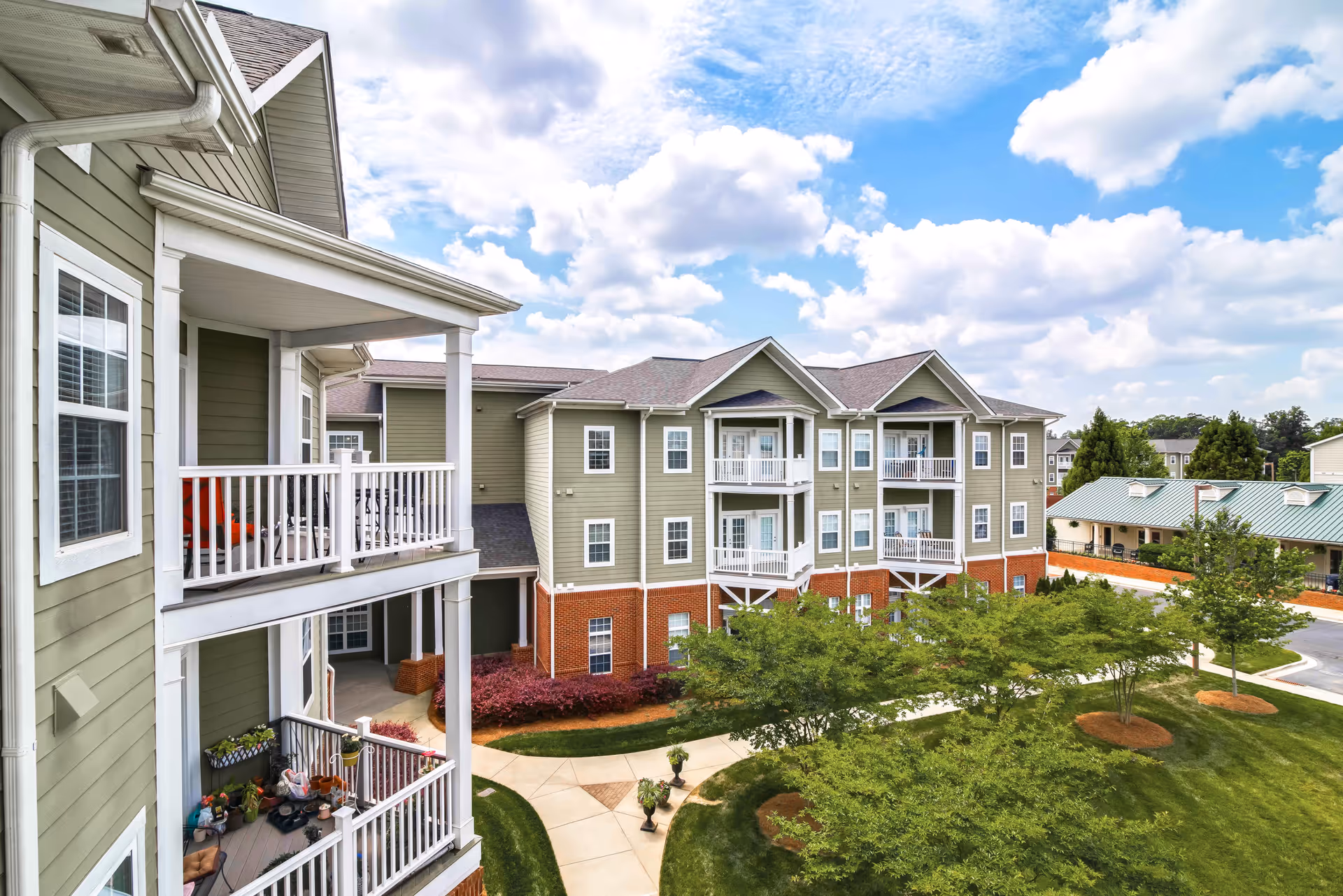 Exterior view of a senior living facility named The Manor showing multiple three-story buildings with green siding and brick bases. The buildings have white balconies with railings, some decorated with plants and chairs. There is a well-maintained lawn with trees and paved walkways under a partly cloudy blue sky.