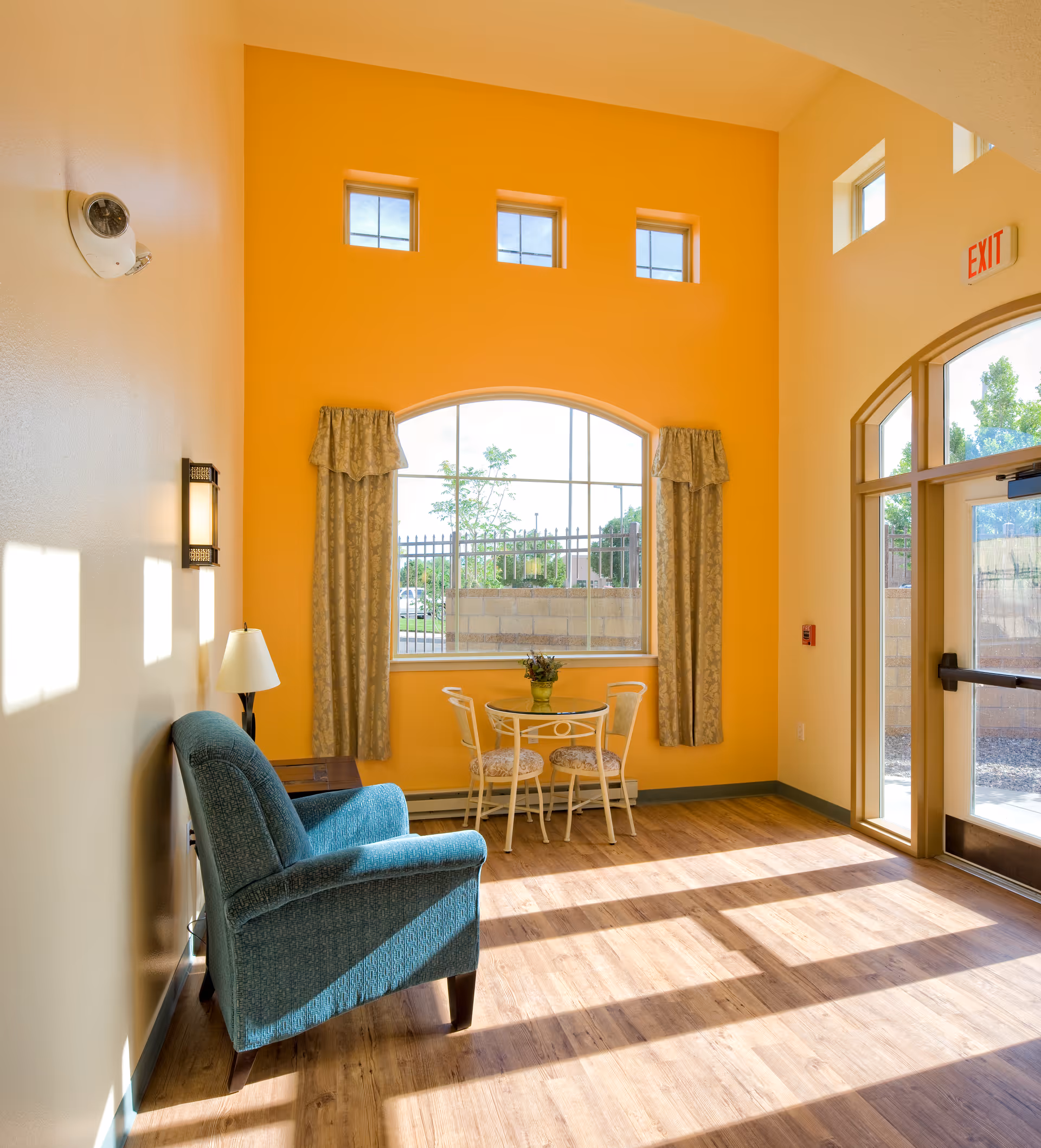Sunlit orange-walled lounge with teal armchair, small round table, and arched window curtains.