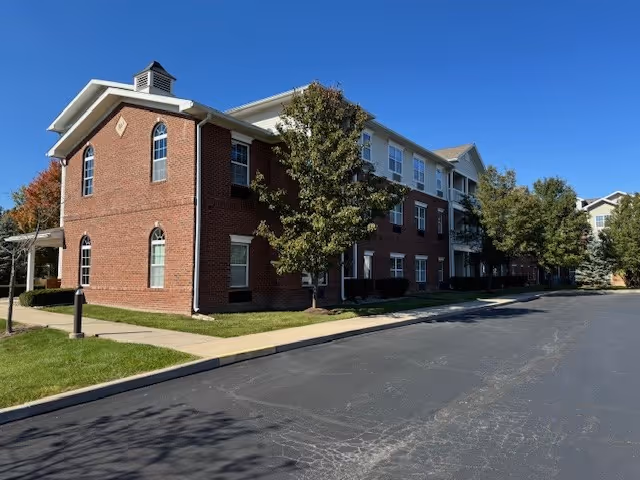 Three-story brick-and-siding senior living building with trees, a sidewalk, and a paved driveway under a clear blue sky.