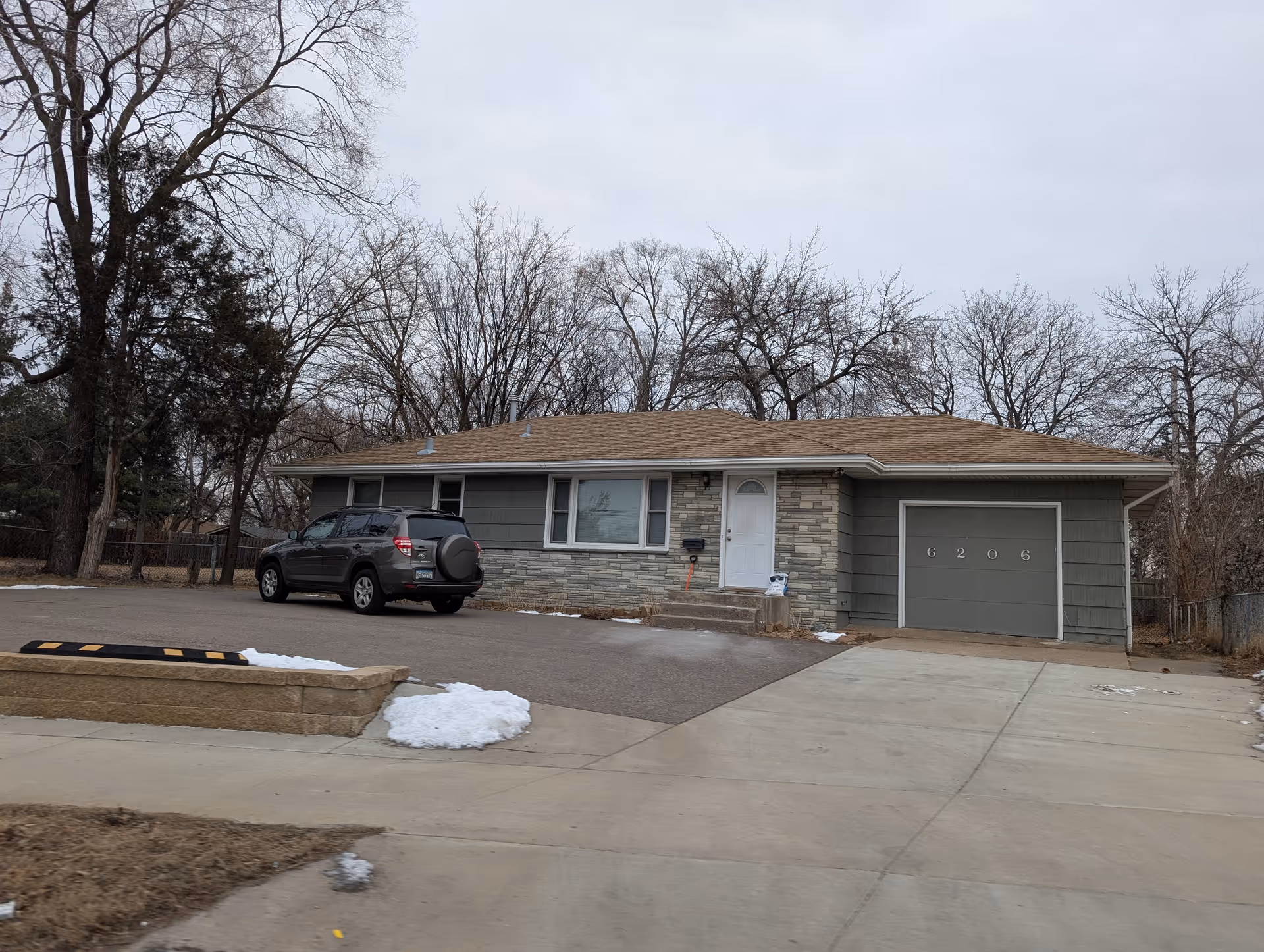 Single-story ranch-style house front with a parked SUV in the driveway, attached garage, and leafless trees behind it.