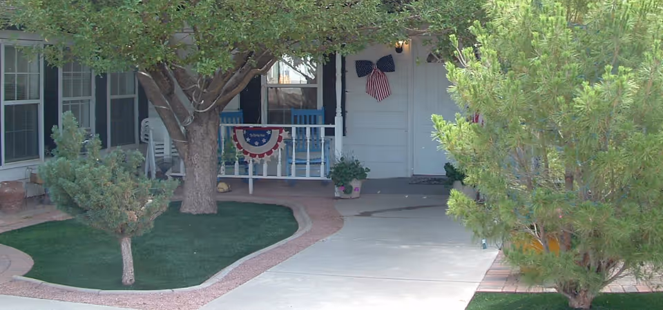 Front entrance of a single-story building showing a porch with white railing, patriotic bunting and flag, small trees and a paved walkway.