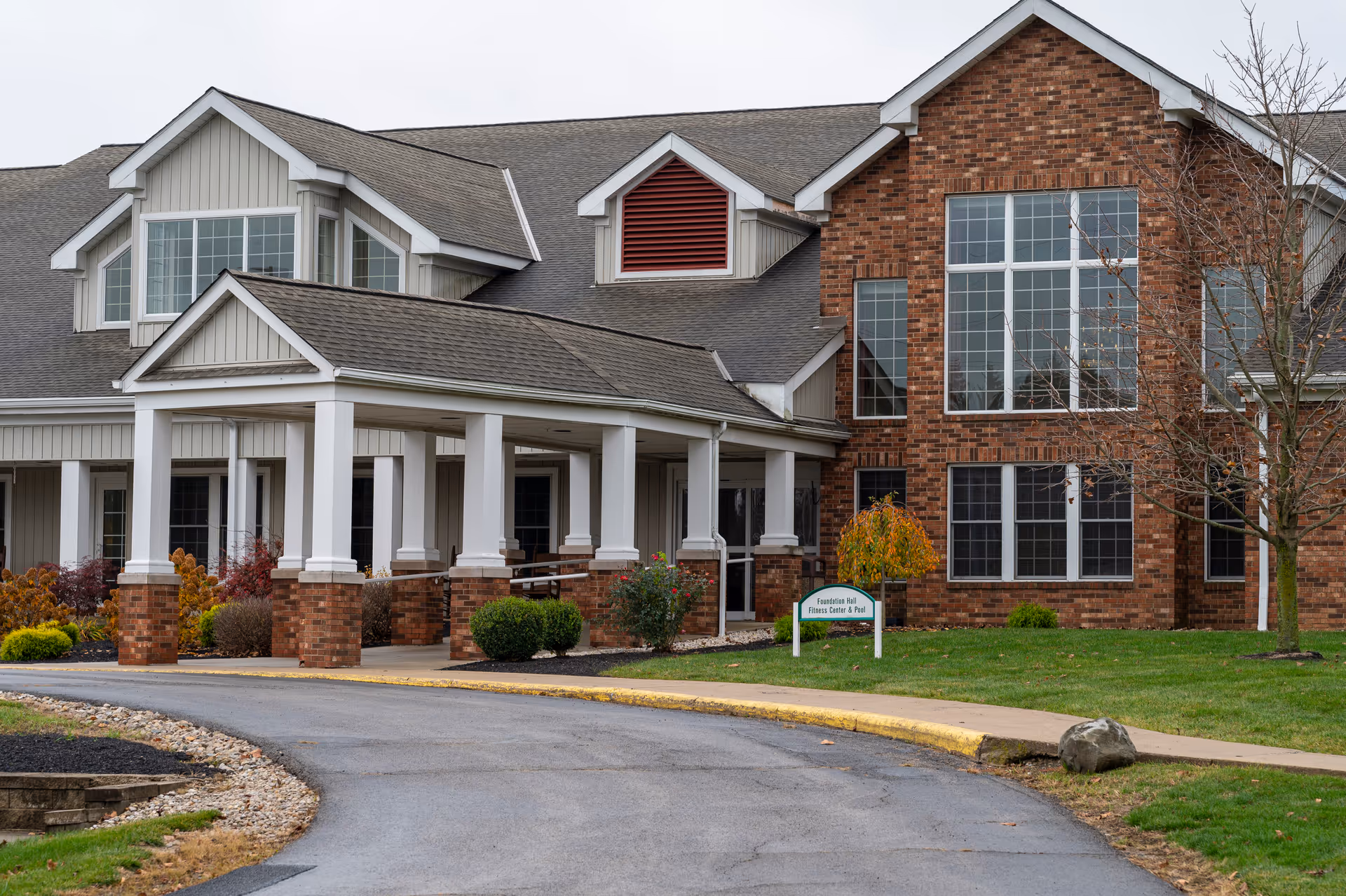 Exterior view of a senior living facility building with a covered entrance supported by white columns and brick bases. The building features large windows, a mix of brick and siding exterior, and a curved driveway leading to the entrance. There is a small sign near the entrance indicating the location of the Fitness Center and Pool.