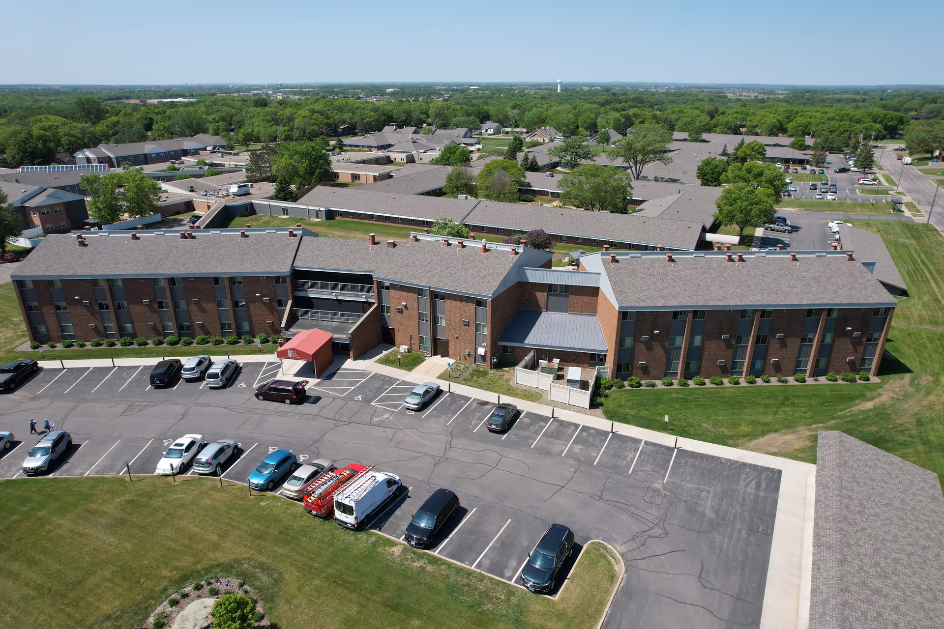 Aerial view of Good Shepherd Community showing a large brick building with multiple windows and a red entrance canopy. The building is surrounded by parking lots with several cars parked and green lawns with trees in the background under a clear sky.