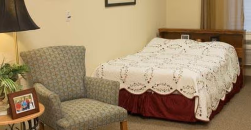 A cozy bedroom in a senior living facility featuring a bed with a white floral quilt and a red bed skirt, a patterned armchair, a small wooden side table with a framed photo and a potted plant, a floor lamp, and a window with blinds.