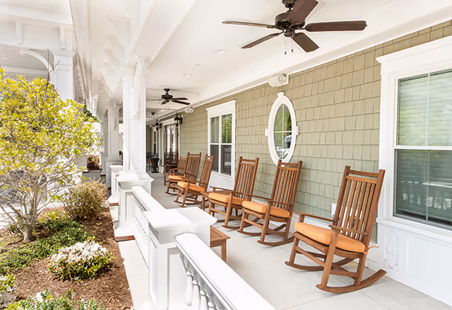 Covered front porch with a row of wooden rocking chairs, ceiling fans, and landscaping at a senior living facility.
