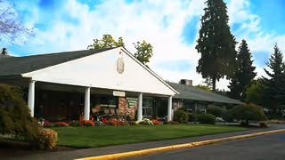Single-story senior living building front with a covered entrance supported by white columns, landscaped lawn, and tall trees.