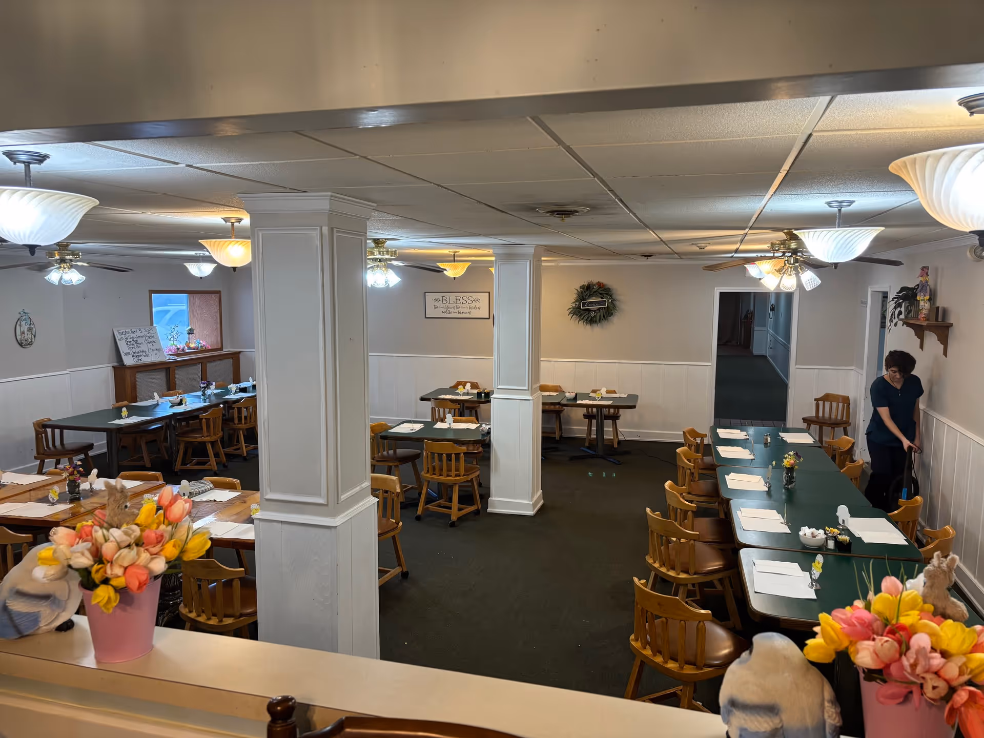 Dining room with multiple green-topped tables and wooden chairs set for meals, central columns, ceiling fans, and a staff member arranging a table.