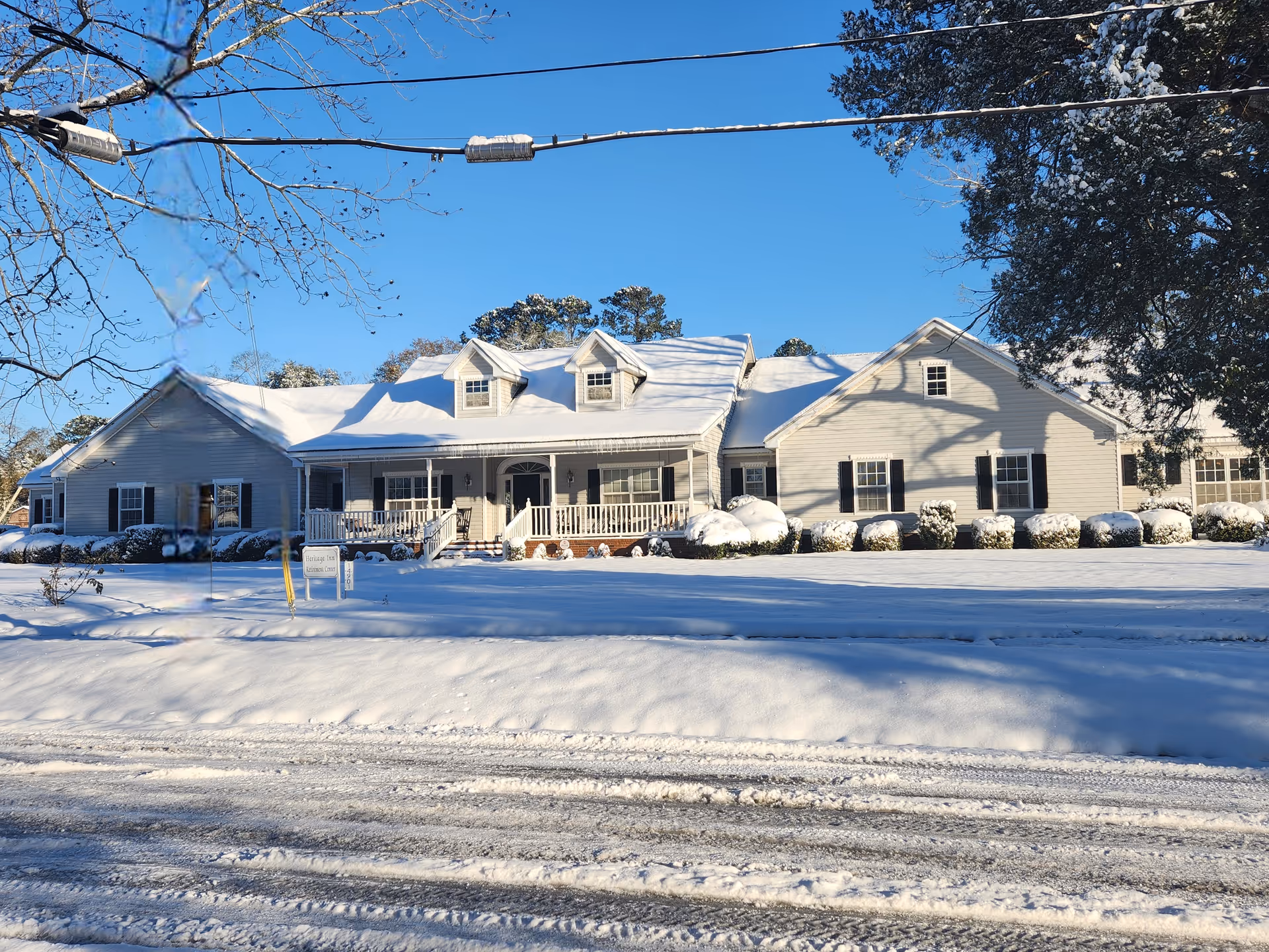 Front view of a single-story white retirement center with a covered porch, snow-covered roof and lawn under a clear blue sky.