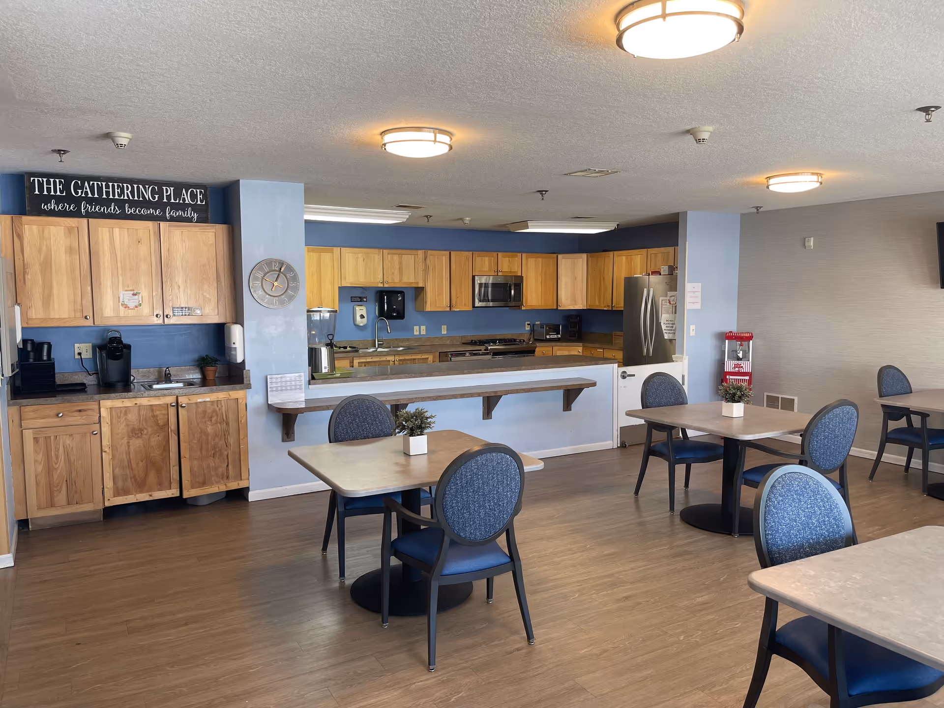A senior living facility dining area with several tables and blue cushioned chairs. The room features a kitchen area with wooden cabinets, a stainless steel refrigerator, microwave, and coffee machines. A sign above the cabinets reads 'THE GATHERING PLACE where friends become family.' The floor is wood, and the walls are painted light blue and beige. Ceiling lights illuminate the space.