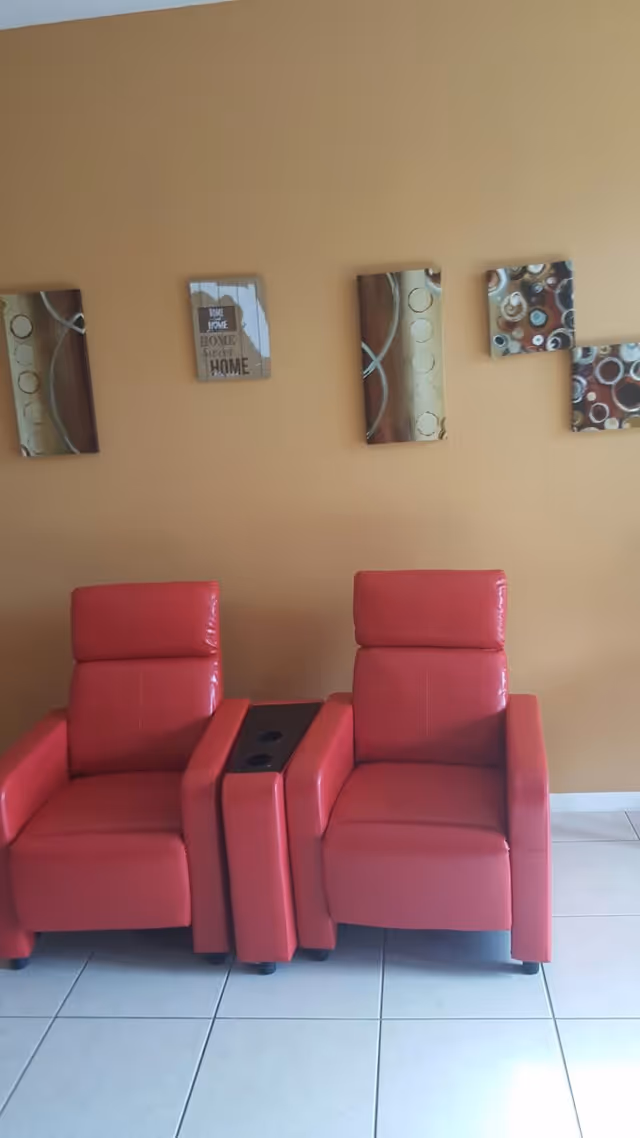 Two red leather recliner chairs with a small matching table with cup holders between them, placed against a beige wall with five decorative wall art pieces featuring abstract and home-themed designs.