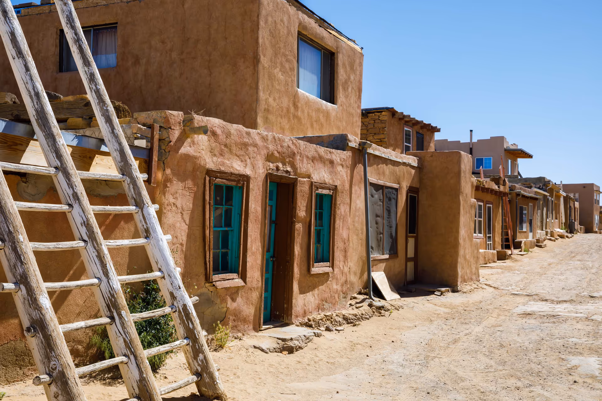 Sunlit adobe houses line a dusty street with ladders propped against walls.