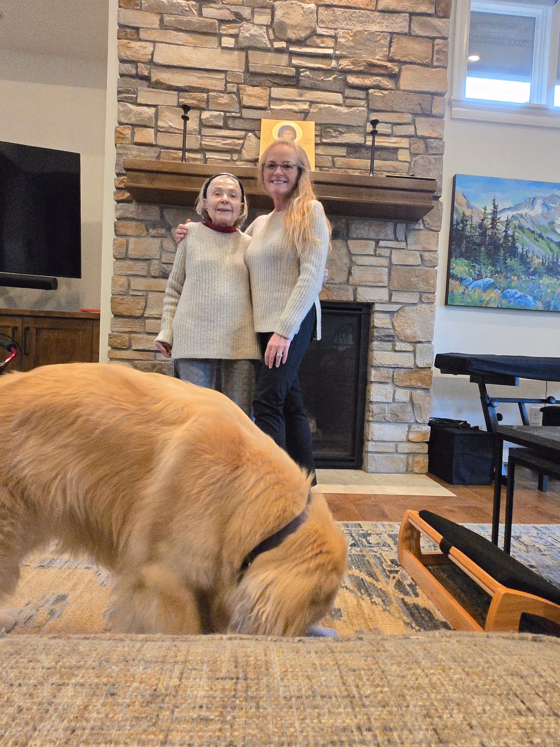Two women stand by a stone fireplace with a large dog in a living room.