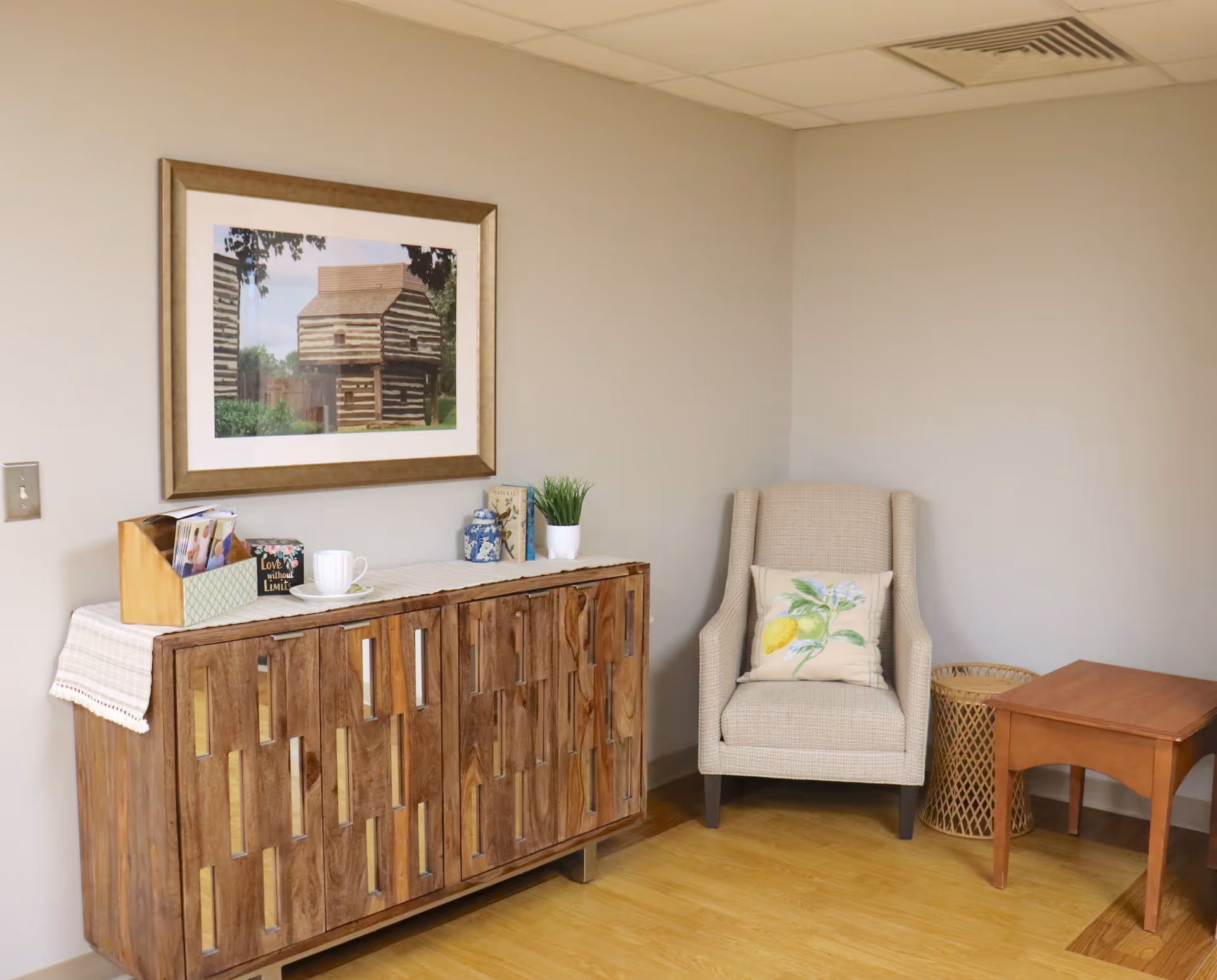 A cozy corner of a room featuring a wooden cabinet with decorative items on top, including a small plant, a cup and saucer, and a framed picture of a log cabin hanging on the wall above. Next to the cabinet is a beige upholstered armchair with a floral cushion, a small wooden side table, and a woven basket on a light wooden floor.