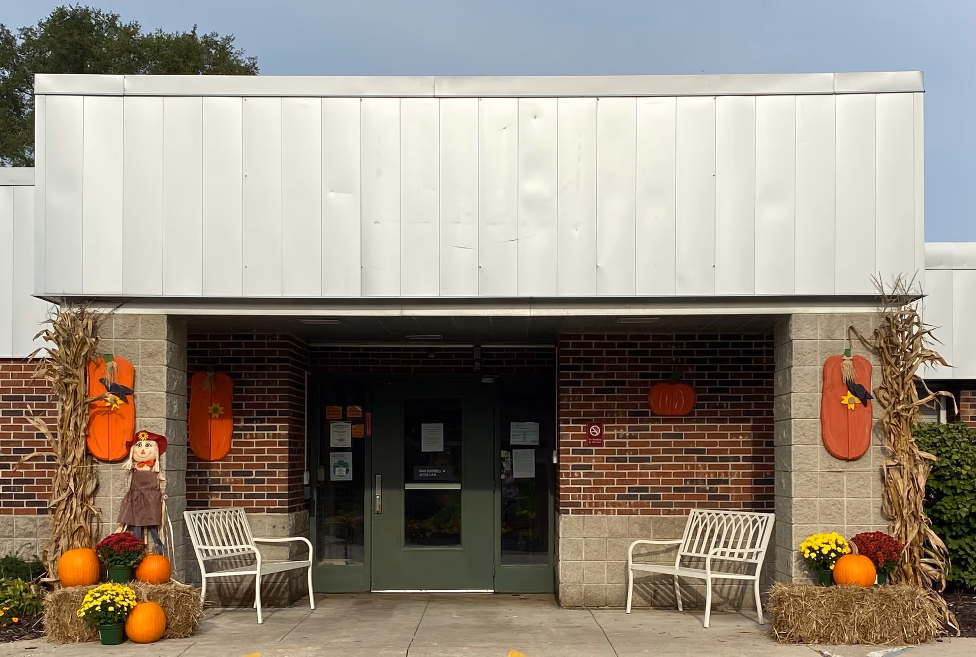 Entrance of a building with double green doors, flanked by two white benches. The entrance is decorated with autumn-themed items including pumpkins, hay bales, yellow and red flowers, cornstalks, and wooden pumpkin decorations on the brick and concrete walls.