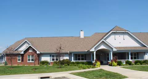 Front exterior view of a single-story senior living facility building with a combination of brick and white siding, a gabled roof, a covered entrance, and a well-maintained lawn under a clear blue sky.