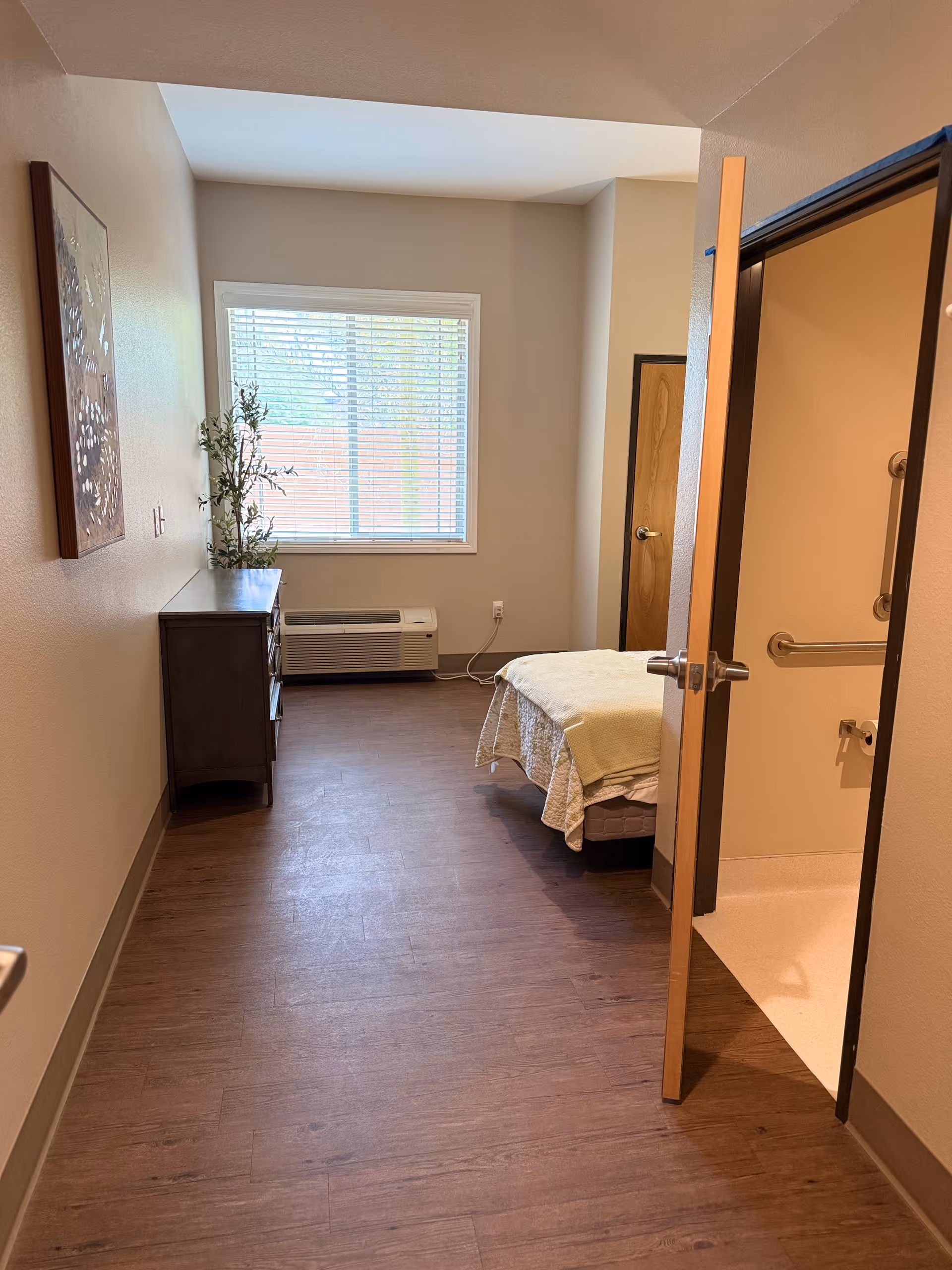 A small bedroom with a single bed covered with a light-colored blanket, a wooden dresser with a plant on top, a window with blinds, and an open door leading to a bathroom with grab bars and a toilet paper holder.