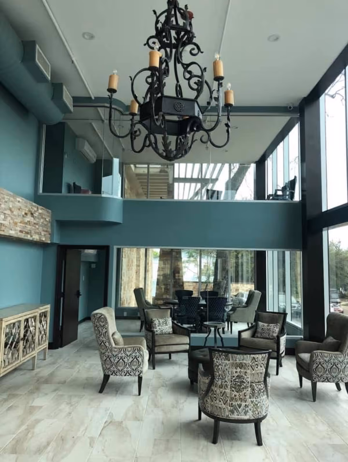 Angled chandelier hangs over a bright lounge in a teal-walled lobby with large windows.