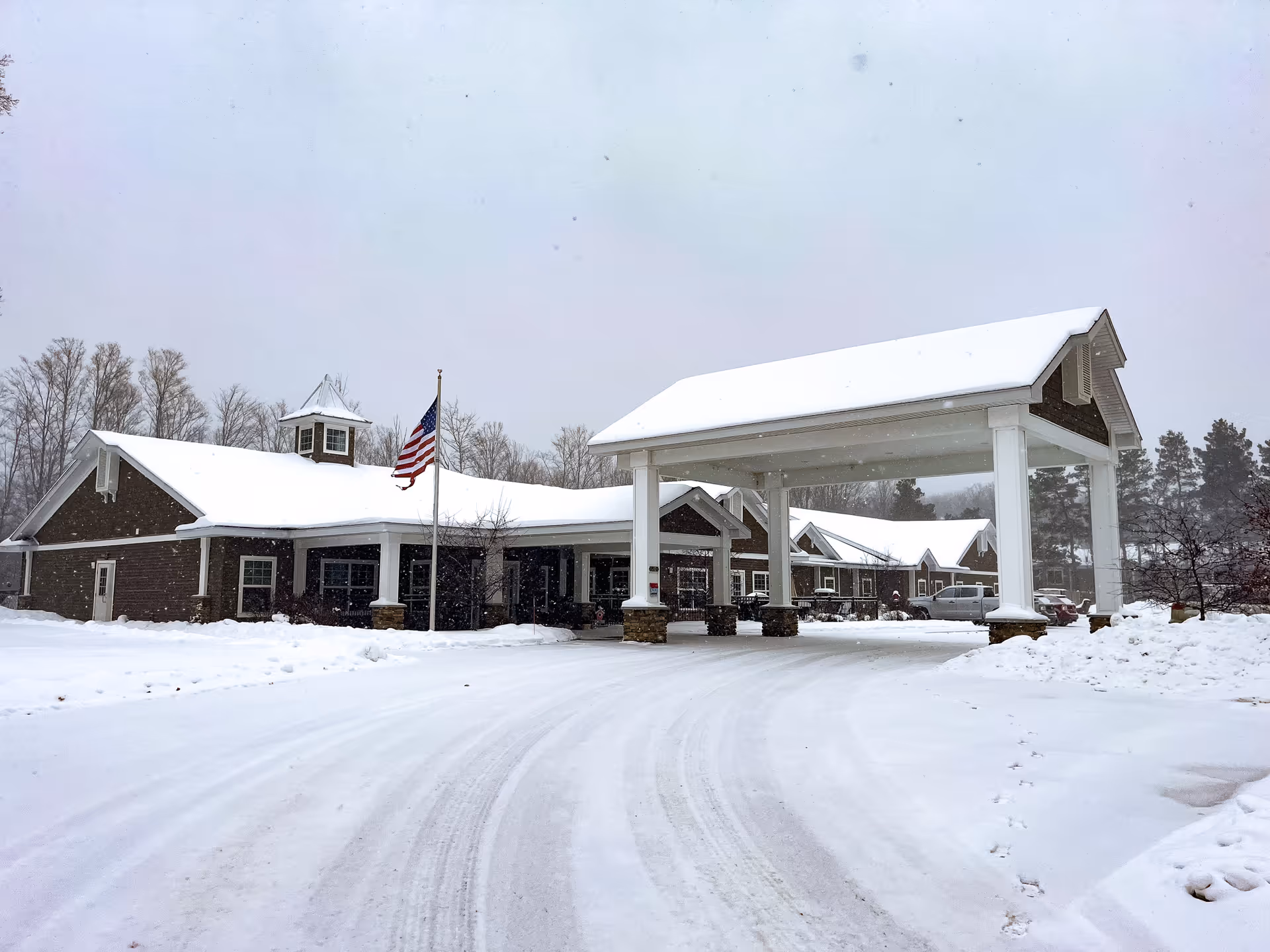 Snow-covered assisted living facility exterior with a large front entrance canopy and American flag.