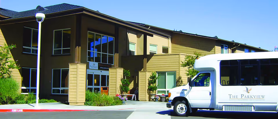 Front entrance of The Parkview senior living building with landscaping and a parked white shuttle van bearing the facility's logo.