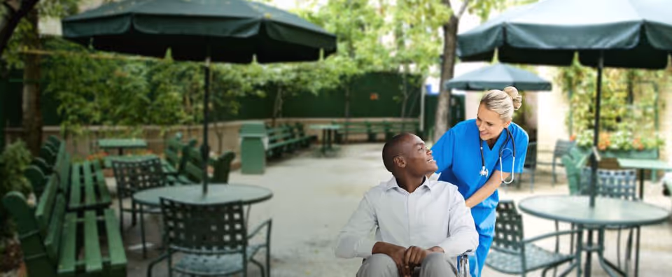 A caregiver in blue scrubs chats with a man in a wheelchair outdoors.