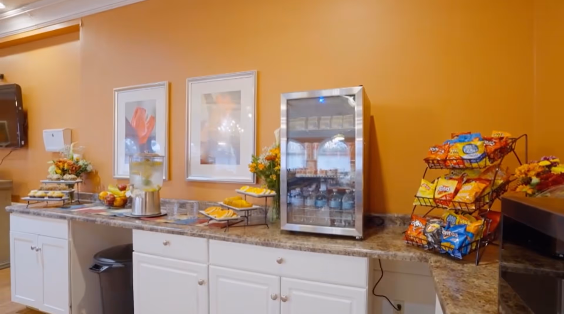 A countertop with a variety of snacks including chips in racks, a small refrigerator stocked with beverages, a water dispenser with lemon slices, plates of food, and floral arrangements against an orange wall with two framed pictures.