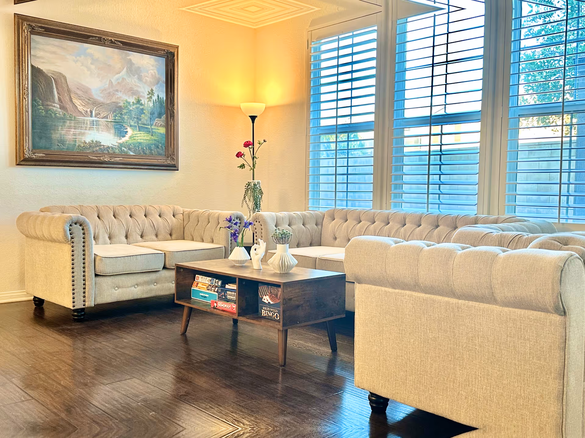 A bright living room with beige tufted sofas arranged around a wooden coffee table holding decorative vases and flowers. Large windows with white blinds allow natural light to fill the room. A floor lamp and a framed landscape painting hang on the wall.